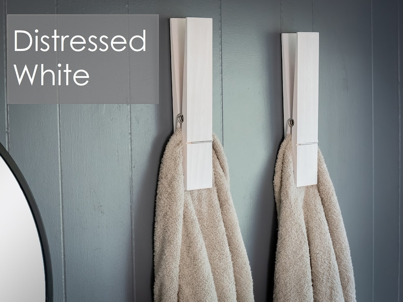 Puede incluir: Dos pinzas de ropa de madera blanca montadas en una pared gris, cada una sujetando una toalla beige. El texto "Distressed White" es visible en la esquina superior izquierda de la imagen.