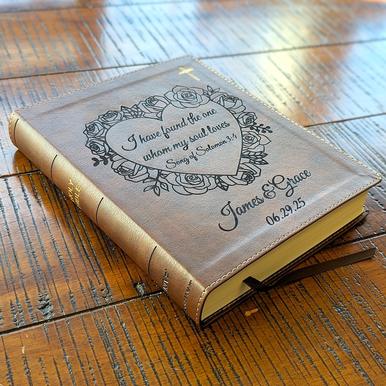Może przedstawiać: Brązowa Biblia w sk&oacute;rzanej oprawie z złotym krzyżem i kwiatowym wzorem na okładce. Na okładce wygrawerowano tekst "I have found the one whom my soul loves Song of Solomon 3:4". Na okładce wygrawerowano r&oacute;wnież imiona "James & Grace" i datę "06.29.25".