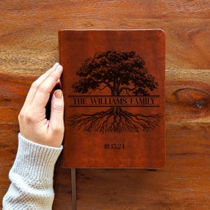 Pode incluir: Um diário de couro castanho com um desenho de árvore e o texto "THE WILLIAMS FAMILY" e "10.15.24". O diário tem uma fita de marcador. A capa é gravada com um gráfico de árvore.