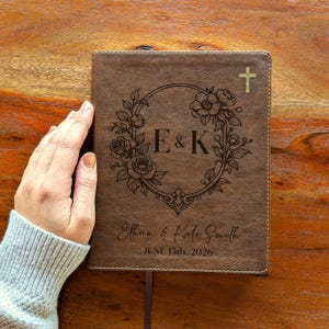 Puede incluir: Diario de cuero marrón con un diseño de corona floral y las iniciales "E & K". Una cruz dorada está en la esquina superior derecha. Los nombres "Ethan & Kate Smith" y la fecha "15 de junio de 2026" están inscritos.