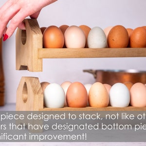 May include: A wooden egg holder with two tiers, each holding a row of eggs. The top tier is being lifted by a hand, showing that the holder is designed to stack. The eggs are a mix of brown and white.