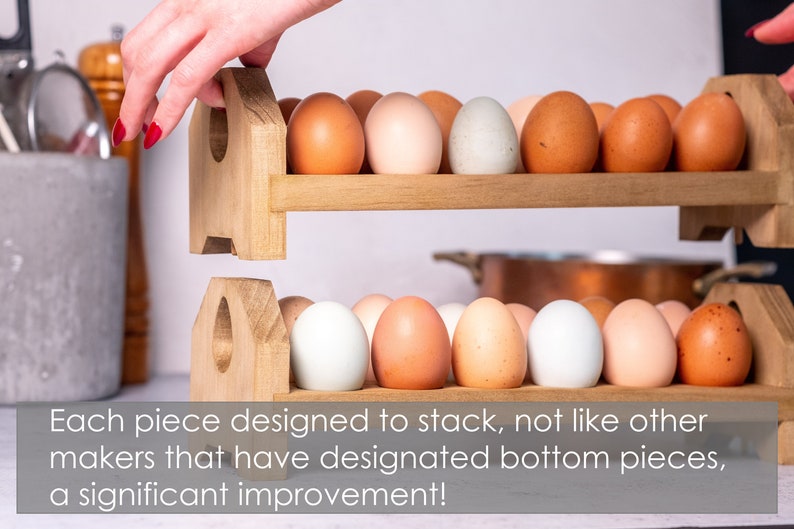 May include: A wooden egg holder with two tiers, each holding a row of eggs. The top tier is being lifted by a hand, showing that the holder is designed to stack. The eggs are a mix of brown and white.