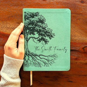 May include: A teal journal featuring a tree design and the words "The Smith Family" in black. It includes a teal bookmark. The journal is held by a person.