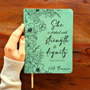 Puede incluir: Un diario verde menta con ilustraciones florales y la frase "She is clothed with strength & dignity". El diario también incluye el texto "Proverbios 31:25" y el nombre "Kate Thompson". Una cinta de marcador beige es visible.