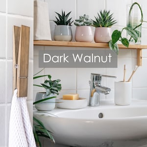 Puede incluir: Un tocador de ba&ntilde;o con un lavabo de porcelana blanca, un grifo cromado y un portacepillos de dientes de cer&aacute;mica blanca. Un toallero de madera con una toalla blanca cuelga de la pared. Una estanter&iacute;a sobre el lavabo contiene tres plantas suculentas en maceta y un jarr&oacute;n de vidrio con una planta trepadora. La estanter&iacute;a est&aacute; etiquetada como "Dark Walnut".