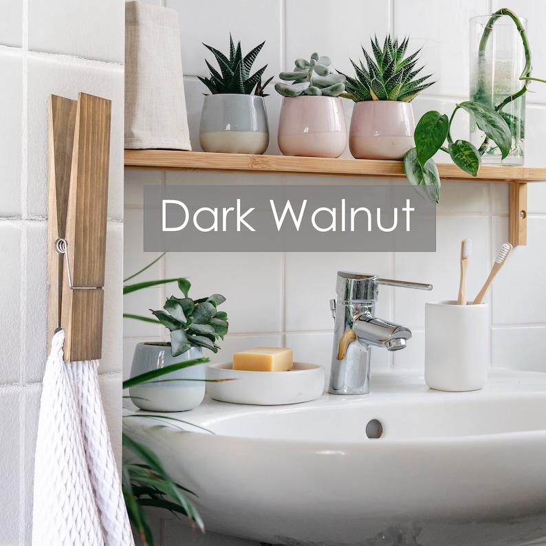 Puede incluir: Un tocador de ba&ntilde;o con un lavabo de porcelana blanca, un grifo cromado y un portacepillos de dientes de cer&aacute;mica blanca. Un toallero de madera con una toalla blanca cuelga de la pared. Una estanter&iacute;a sobre el lavabo contiene tres plantas suculentas en maceta y un jarr&oacute;n de vidrio con una planta trepadora. La estanter&iacute;a est&aacute; etiquetada como "Dark Walnut".