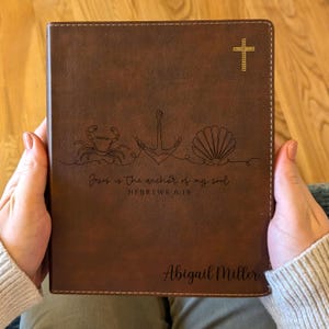 Könnte beinhalten: Braunes Lederbibel mit einem goldenen Kreuz und nautischer Strichzeichnung einer Krabbe, eines Ankers und einer Muschel. Der Text "Jesus is the anchor of my soul, Hebrews 6:19" ist eingraviert, zusammen mit dem Namen "Abigail Miller".