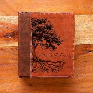 May include: A brown leather journal featuring a tree design and the text "The Smith Family." The tree illustration is dark brown and covers most of the journal's front. The spine is a darker brown with white stitching.
