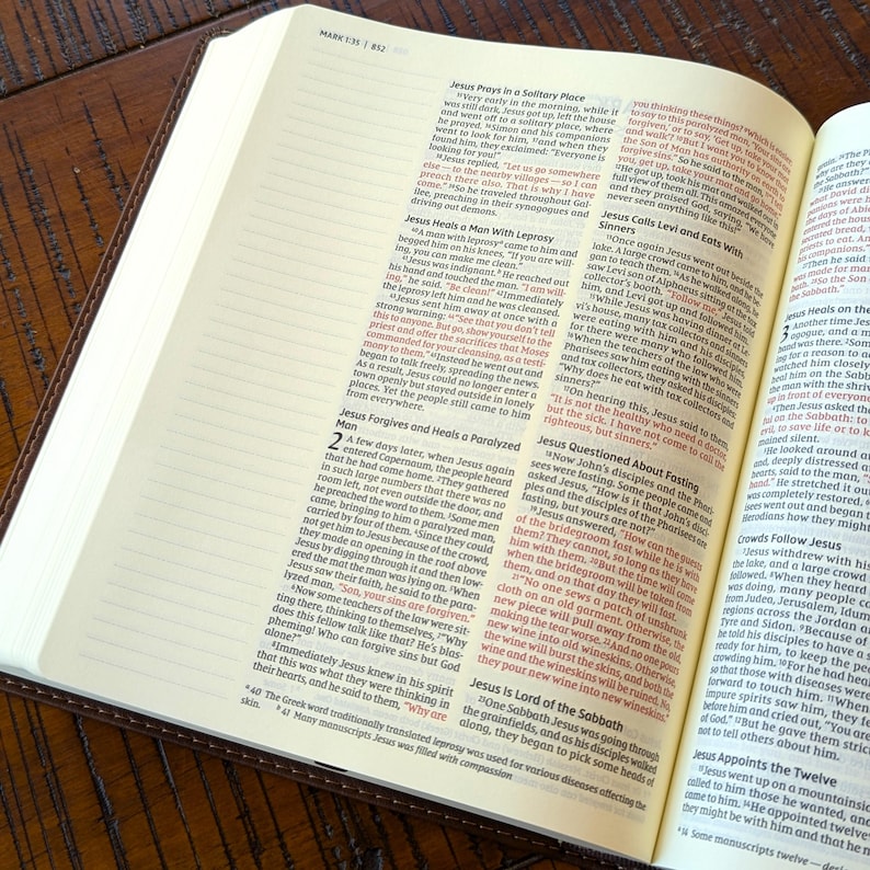 Może przedstawiać: Zbliżenie na brązową, sk&oacute;rzaną Biblię otwartą na Księgę Marka, rozdział 1, wersety 35-45. Tekst jest podświetlony na czerwono i opowiada o uzdrowieniu trędowatego przez Jezusa oraz o przebaczeniu i uzdrowieniu sparaliżowanego mężczyzny.