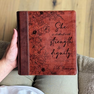 May include: A brown leather Bible cover with floral designs and the words "She is clothed with strength & dignity" in elegant script. The cover also includes the name "Kate Thompson" and the Bible verse Proverbs 31:25.