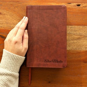 May include: A brown leather journal with the name "Alice Martin" embossed on the cover. The journal has a red ribbon bookmark and is on a wooden surface.