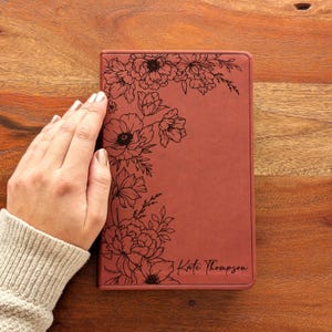 Op de afbeelding: Een terracotta-kleurig notitieboek met een bloemenontwerp in zwarte inkt. De bloemen zijn geëtst in de linkerbovenhoek en de naam "Kate Thompson" is in script in de rechterbenedenhoek geschreven.