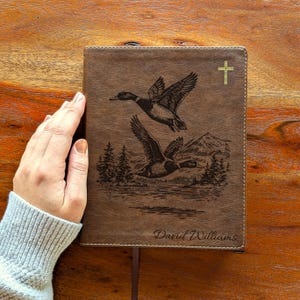 May include: Brown leather-bound journal with a detailed engraving of two ducks in flight over a landscape with trees and mountains. A gold cross is in the upper right corner, and the name "David Williams" is at the bottom.