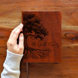 May include: A brown leather journal featuring a tree design and the text "The Smith Family". The tree illustration is dark brown. The journal is held by a hand. The journal has a ribbon bookmark.