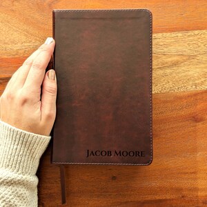 Könnte beinhalten: Ein dunkelbraunes, ledergebundenes Notizbuch mit dem Namen "JACOB MOORE", der in der unteren rechten Ecke eingeprägt ist. Das Notizbuch hat ein Leseband und wird von einer Person gehalten, die einen hellen Pullover trägt. Das Notizbuch liegt auf einer Holzoberfläche.