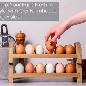 May include: A two-tiered wooden egg holder with brown eggs and blue eggs on each shelf. The egg holder is on a white countertop with a gray concrete utensil holder and a brown pot in the background. The text "Keep Your Eggs Fresh in Style with Our Farmhouse Egg Holder!" is at the top of the image.