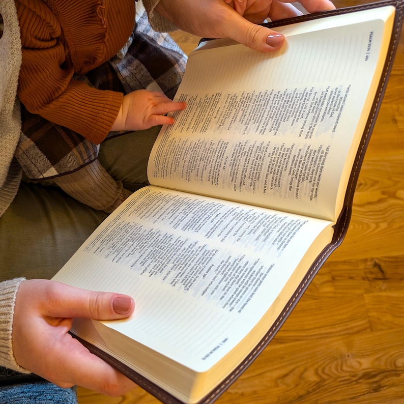 Może przedstawiać: Książka w brązowej sk&oacute;rzanej oprawie z białymi stronami i ręką trzymającą ją otwartą. Książka jest otwarta na stronie z małym czarnym tekstem.
