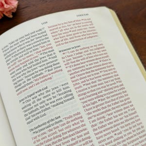 May include: An open Bible with text from John 5:40, highlighted in red. The pages are cream-coloured, set against a brown wooden surface. The book is open to a specific passage.