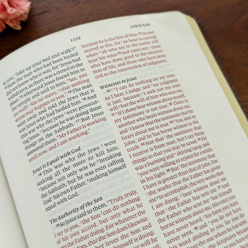May include: An open Bible with text from John 5:40, highlighted in red. The pages are cream-coloured, set against a brown wooden surface. The book is open to a specific passage.