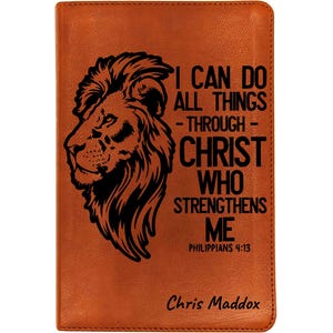 May include: A brown leather journal with a lion's head design and the text "I CAN DO ALL THINGS - THROUGH - CHRIST WHO STRENGTHENS ME" and "PHILIPPIANS 4:13". The name "Chris Maddox" is at the bottom.