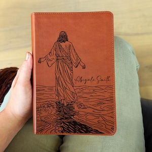 Op de afbeelding: Een bruin leren boek met een gegraveerde afbeelding van een persoon die over water loopt. De figuur is van achteren te zien, met open armen. De naam "Abigale Smith" staat eronder geschreven.