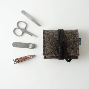 May include: A gray felt pouch with a black leather strap and the text "manula.k" printed on the side. The pouch contains a set of manicure tools, including a nail clipper, tweezers, nail file, and scissors.