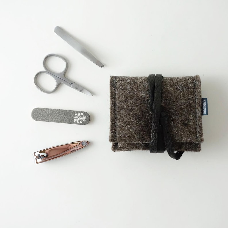 May include: A gray felt pouch with a black leather strap and the text "manula.k" printed on the side. The pouch contains a set of manicure tools, including a nail clipper, tweezers, nail file, and scissors.