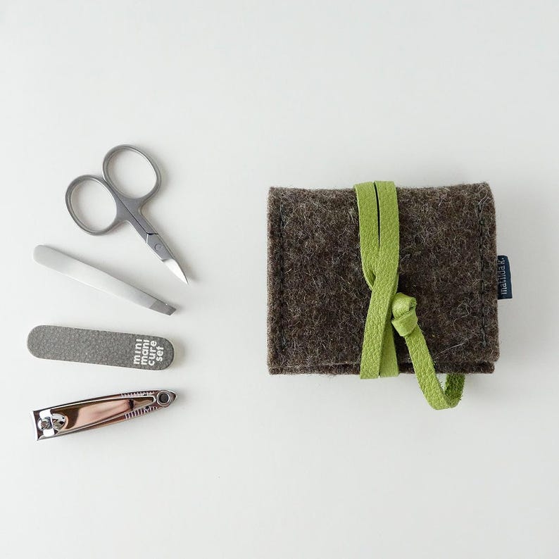 May include: A brown felt pouch with a green strap, containing a mini manicure set. The set includes a nail clipper, nail file, tweezers, and small scissors.