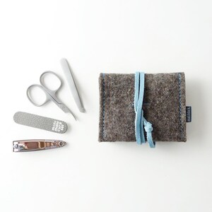 May include: A gray felt pouch with a blue leather strap and the word "Feltbag" printed on it. The pouch contains a set of manicure tools, including a nail file, nail clippers, and tweezers.