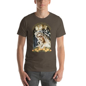 May include: A brown t-shirt with a religious image of a woman in a crown and veil, framed by yellow roses. The text "AVE MARIA GRATIA PLENA" arches above the image in gold. The shirt is worn by a person.