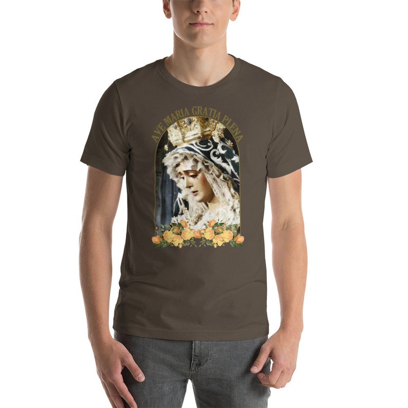 May include: A brown t-shirt with a religious image of a woman in a crown and veil, framed by yellow roses. The text "AVE MARIA GRATIA PLENA" arches above the image in gold. The shirt is worn by a person.