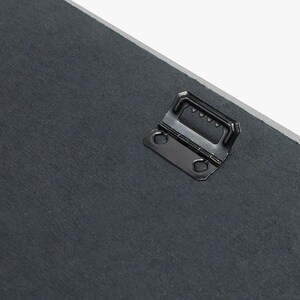 Puede incluir: Un gancho de metal negro para colgar con un dise&ntilde;o de dientes de sierra en la parte superior y dos agujeros en los lados. El gancho est&aacute; unido a una superficie gris oscuro.