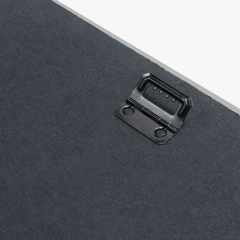 Puede incluir: Un gancho de metal negro para colgar con un dise&ntilde;o de dientes de sierra en la parte superior y dos agujeros en los lados. El gancho est&aacute; unido a una superficie gris oscuro.