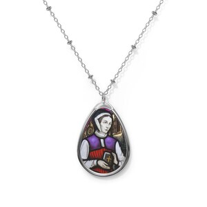 Puede incluir: Collar de cadena de plata con un colgante en forma de lágrima que presenta una imagen de vitral de una mujer con una túnica morada y roja que sostiene un libro.