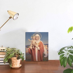May include: A religious art print featuring a woman in a red robe holding a child. Both figures have halos. The background depicts a landscape with buildings under a blue sky. The print is displayed on a wooden surface.