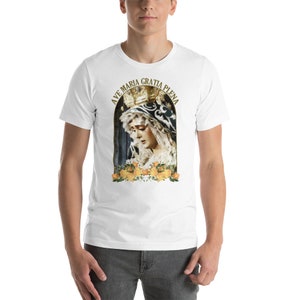 May include: White t-shirt with a religious image of a crowned woman, framed by yellow roses. The text "AVE MARIA GRATIA PLENA" is arched above the image in gold lettering. The shirt is made of a soft material.