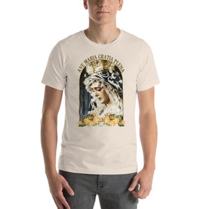 May include: A cream-coloured t-shirt with a religious image of a woman wearing a crown, framed by yellow roses. The text "AVE MARIA GRATIA PLENA" is arched above the image. The shirt is made of a soft material.
