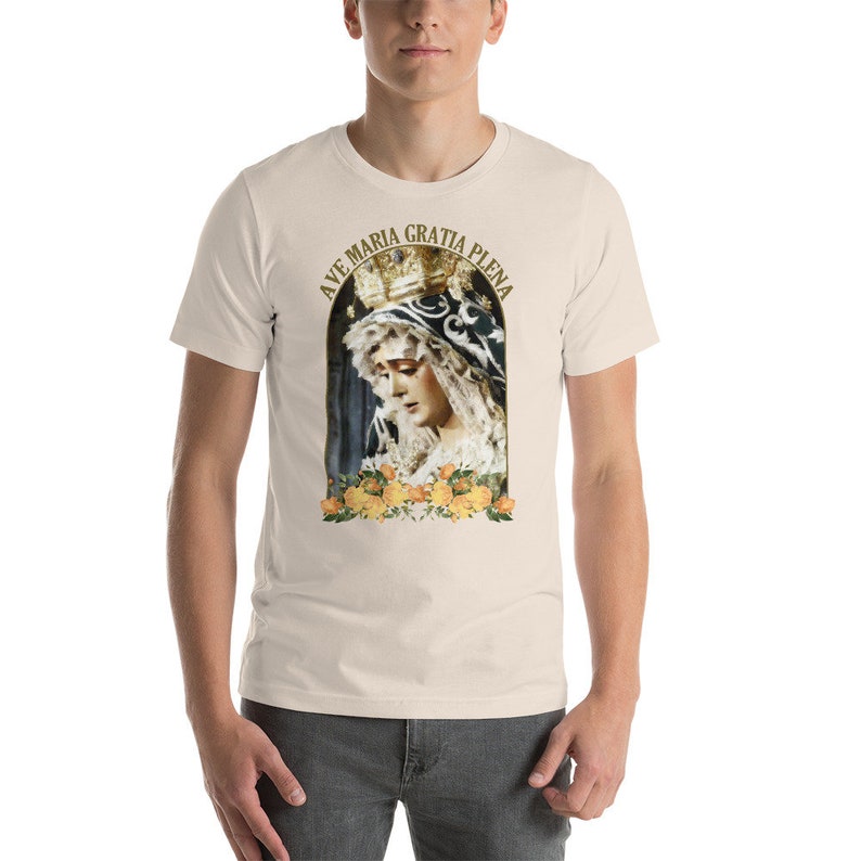May include: A cream-coloured t-shirt with a religious image of a woman wearing a crown, framed by yellow roses. The text "AVE MARIA GRATIA PLENA" is arched above the image. The shirt is made of a soft material.