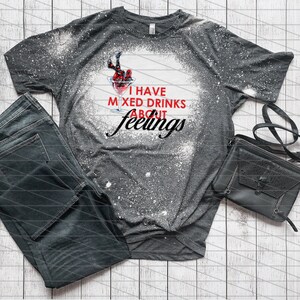 May include: A gray t-shirt with a graphic of a cocktail and the text "I HAVE MIXED DRINKS ABOUT feelings". The shirt is paired with dark wash jeans and a black purse.