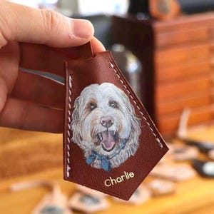 May include: A brown leather tag with a painted dog portrait. The dog has white fur and a blue bow tie. The name "Charlie" is printed in white. The tag is stitched with white thread around the edges.