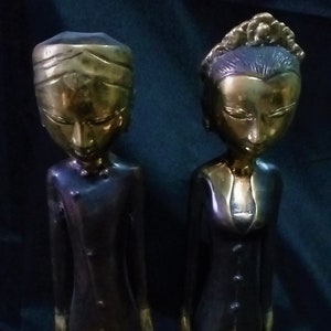 Puede incluir: Dos estatuas de bronce de figuras con vestimenta tradicional indonesia. Las figuras tienen rasgos alargados y se representan de pie. Las estatuas tienen una combinación de colores oro y negro, con detalles intrincados en la ropa y los tocados.