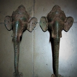May include: A pair of antique bronze elephant head door handles. Each handle depicts an elephant head with detailed ears, a long trunk, and a decorative headdress. The handles are mounted on a matching base and have a weathered, aged appearance.