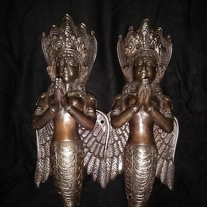 Può includere: Coppia di maniglie per porte color bronzo raffiguranti figure con le mani giunte in preghiera. Le figure hanno copricapi ornati, ali piumate e corpi inferiori squamosi. Le maniglie sono montate su uno sfondo scuro.