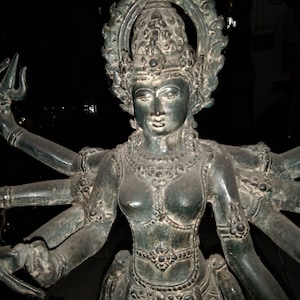 May include: A dark bronze statue of a multi-armed deity, possibly a goddess. The statue has a serene facial expression, ornate headdress, and detailed clothing. The arms are gracefully positioned, holding various objects.