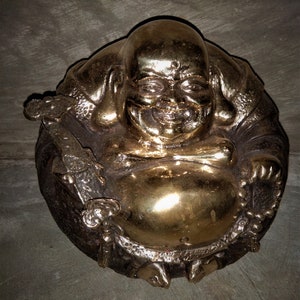 Puede incluir: Una estatua de Buda sonriente de color bronce. La figura está representada en posición sentada con una gran barriga y una cara sonriente. La estatua tiene un acabado liso y pulido en la barriga y la cabeza, con detalles texturizados en las túnicas.