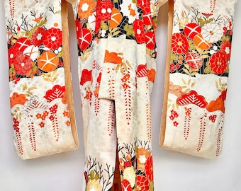 May include: Cream-coloured kimono with vibrant floral patterns in red, orange, and white. The traditional Japanese garment features long sleeves and a detailed design, displayed on a wooden stand. The kimono's design includes black accents.