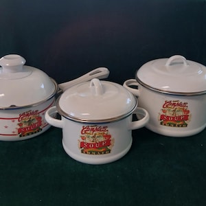 May include: Three white enamel soup pots with red and white Campbell's Soup labels. The pots have black rims and handles. The labels feature the Campbell's Soup logo and the words "Soup" and "Tomato".