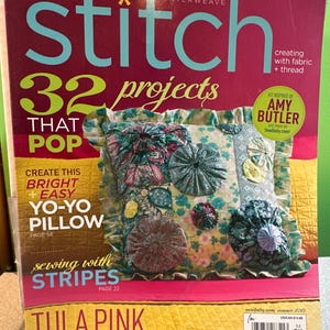 Op de afbeelding: Een tijdschriftomslag voor "stitch" met een kleurrijk kussen met bloemmotieven. De omslag bevat tekst als "32 projects that pop" en "sewing with stripes". Het tijdschrift bevat ook "Tula Pink".
