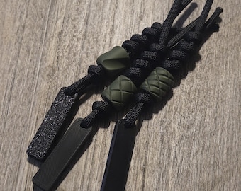 May include: Three black paracord zipper pulls with olive green accents. Each pull features a different design: a textured rectangle, a smooth rectangle, and a textured bead. The pulls are knotted at the top.