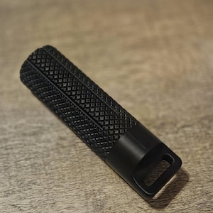 Op de afbeelding: Een zwart, rechthoekig metalen object met een textuur, diamantpatroon grip. Het object heeft een glad, cilindrisch uiteinde met een kleine, rechthoekige opening. Het item ligt op een houten oppervlak.